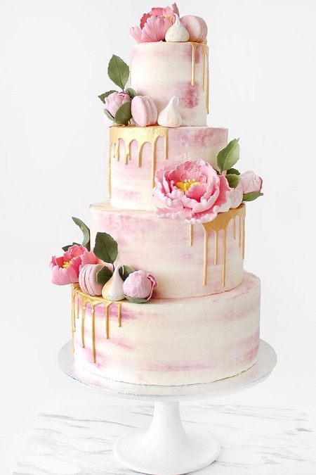 In questa foto, una torta che cola a più piani con glassa bianca, rosa e dorata e dettagli a forma di fiori, meringhe, macaron nei toni del bianco e del rosa brillante 