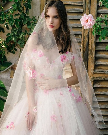 In questa foto, una modella indossa un abito da sposa bianco in tulle con fiori rosa brillante, il colore della Biodiversità di Pantone 