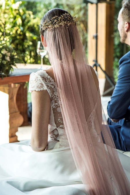 In questa foto, una sposa seduta, fotografata da dietro, indossa un velo rosa attaccato a una coroncina di fiori 