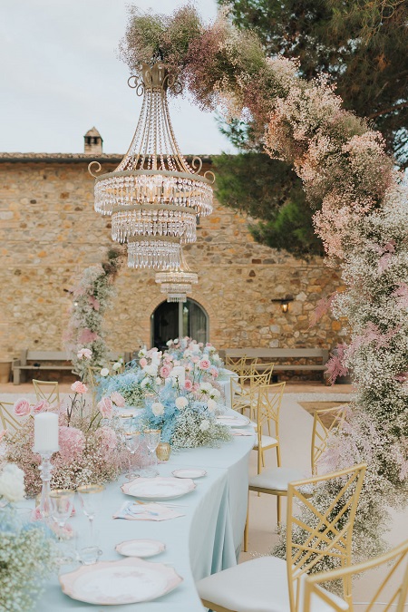 In questa foto, l'allestimento di un tavolo di nozze all'esterno con tovaglia azzurra, composizioni di fiori azzurri e rosa sul tavolo e un semi arco floreale da cui pende un lampadario sospeso con cristalli 