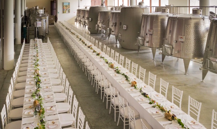 In questa foto due grandi tavoli imperiali addobbati per ospitare un ricevimento di nozze, posizionati all'interno della sala di produzione del vino de Le Palaie, in Toscana