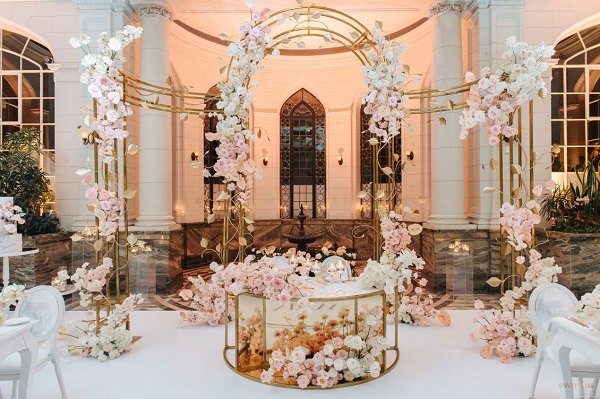 In questa foto, il tavolo degli sposi è decorato con archi dorati e fiori bianchi e rosa brillante, il colore della Biodiversità di Pantone