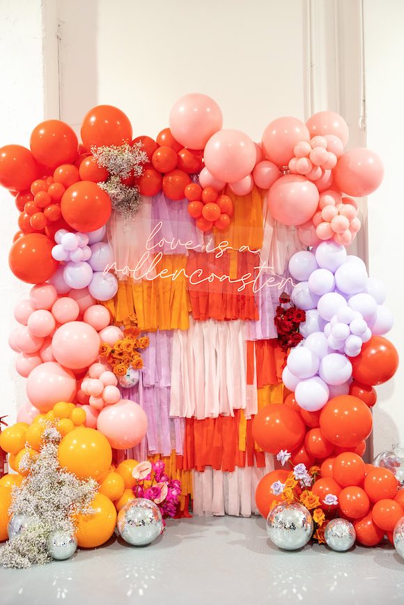 In questa foto un backdrop con palloncini e nastri iper colorati, nei toni dell'arancione chiaro e scuro, rosa e lilla. Appesa sul backdrop, la scritta neon "Love is a rollercoaster"