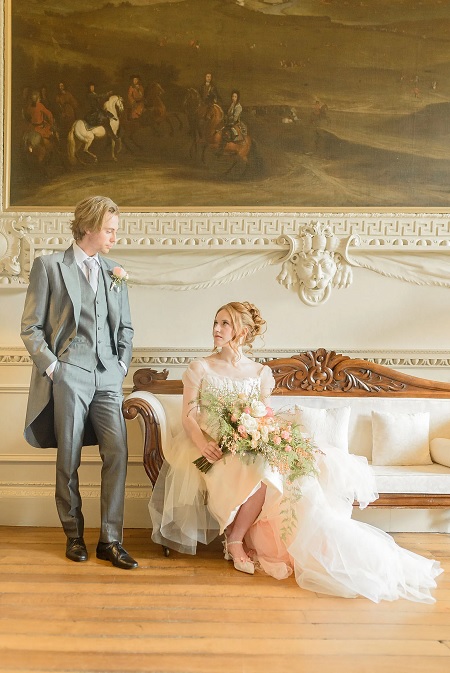 In questa foto, una coppia di sposi, lei è seduta su un divano, indossa un abito bianco e tiene in mano un bouquet, lui è in piedi con un tight grigio