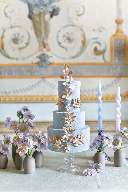 In questa foto, una torta nuziale a tre piani azzurro polvere con al centro un lungo ramo di fiori bianchi e foglie dorate