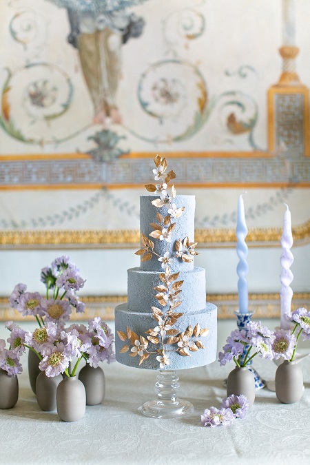 In questa foto, una torta nuziale a tre piani azzurro polvere con al centro un lungo ramo di fiori bianchi e foglie dorate
