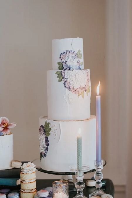 In questa foto, una torta matrimonio stile Bridgerton a tre piani con base bianca e fiori viola e lilla disegnati con la pasta di zucchero 