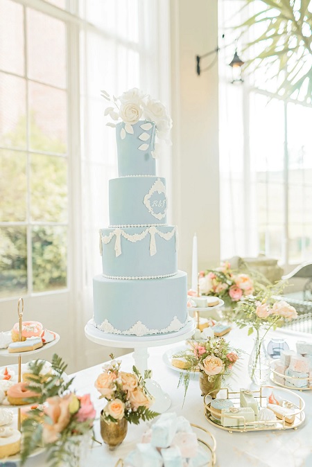 In questa foto, un tavolo dolci con una torta nuziale a quattro piani con base azzurro polvere e decori di pizzo, perline, iniziali degli sposi e cake topper a forma di rose in bianco