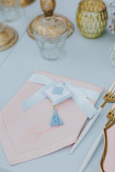In questa foto, il particolare di un tavolo apparecchiato con una tovaglia azzurro chiaro e un tovagliolo rosa piegato a triangolo, tenuto fermo da un nastro azzurro e una piccola decorazione a forma di cuscino bianco e azzurro con ciondolo a nappa azzurro