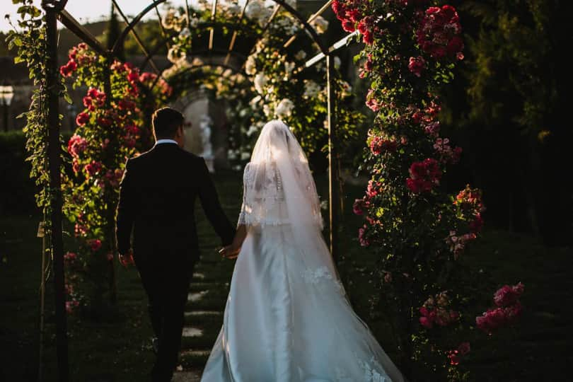 In questa foto due sposi di spalle attraversano l'arco di rose che si trova in uno dei giardini di Villa Scorzi, location per matrimoni in Toscana, nella zona di Pisa