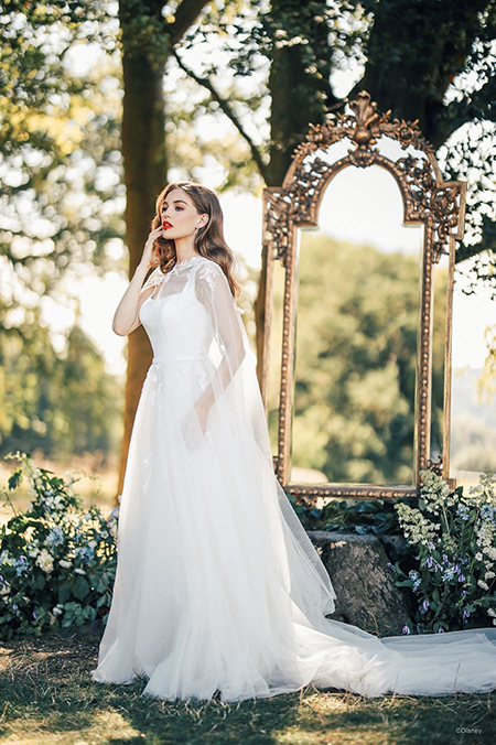 In questa foto la modella indossa un abito da sposa disney modello Snow White con scollo quadrato e gonna di tulle con fiori con mantella