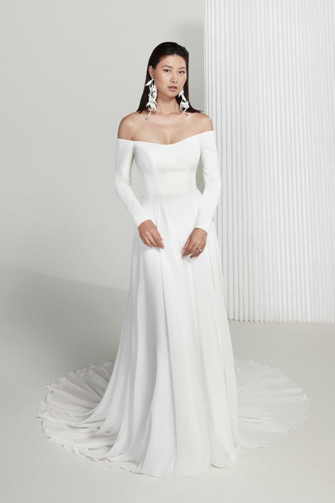 In questa foto la modella indossa un abito da sposa semplice 2023 con spalle scoperte e maniche lunghe justin alexander