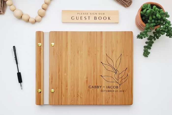 In questa foto un guestbook con copertina in legno su cui sono incisi i nomi degli spos, la data del matrimonio e un ramoscello di foglie