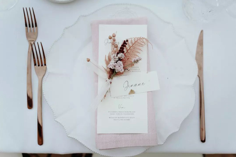 In questa foto una mise en place elegante con posate di colore oro, piatto in porcellana bianca, tovagliolo in stoffa di colore rosa, fiori di campo  abbinati e un segnaposto in carta cotone