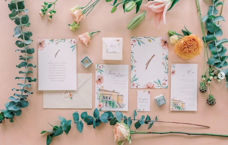 In questa foto un coordinato di matrimonio romantico con disegni di foglie e fiori di colore rosa e uno che rappresenta la location delle nozze realizzata con acquerelli