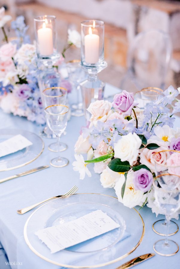 In questa foto il dettaglio di un mise en place per un matrimonio in stile Bridgerton. In primo piano è inquadrato un piatto in vetro trasparente con bordi colore oro abbinato a bicchieri e posate. Sul piatto è posizionato un menù. Al centro del tavolo sono presenti composizioni di fiori di colore bianco, rosa e lilla. Il tovagliato è di colore azzurro polvere