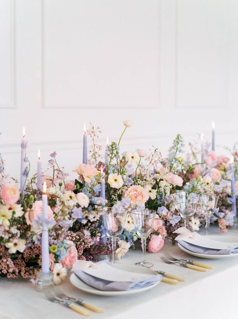 In questa foto un allestimento floreale per matrimonio in stile Bridgerton con fiori di colore rosa, lilla, azzurri su un tavolo rettangolare. L'allestimento è completato da candele accese di colore lilla