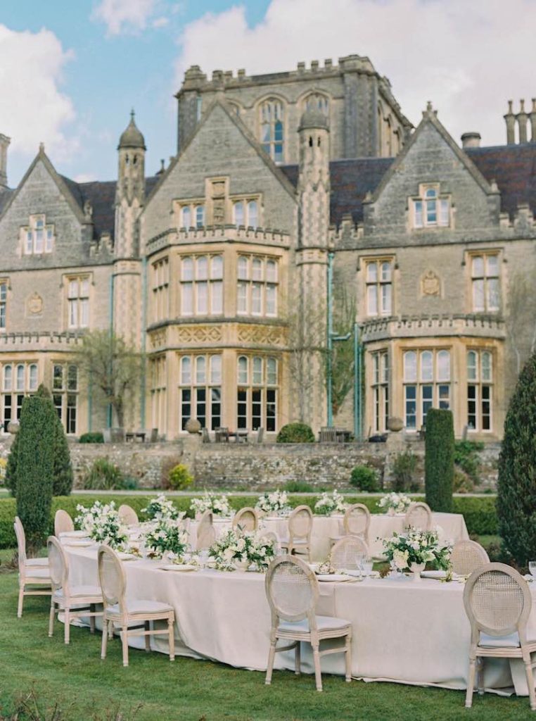 In questa foto un allestimento di nozze in stile Bridgerton sul giardino di un castello che viene mostrato sullo sfondo. In primo piano sono inquadrati i tavoli del matrimonio decorati con tovagliato di colore rosa e fiori di colore bianco, abbinati a sedie eleganti con schienale rotondo 