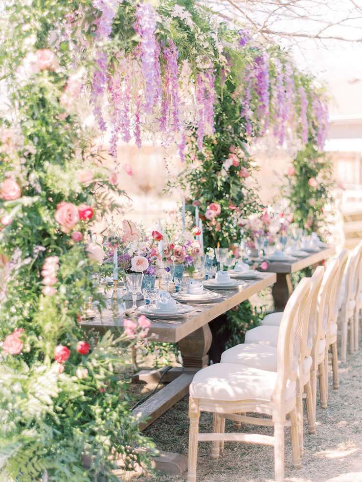 In questa foto l'allestimento di un matrimonio in stile Bridgerton con tavoli imperiali, fiori a casacata sui tavoli e al centro tavolo, sedie classiche colore avorio. I fiori usati sono di colore lilla, rosa e fucsia