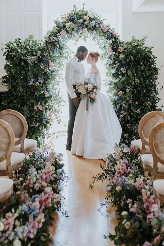 In questa foto due sposi che si guardo di un matrimonio in stile Bridgerton in piedi davanti ad un arco di fiori di colore rosa e turchese. La sposa tiene un bouquet con fiori abbinati. Gli stessi fiori sono presenti lungo il corridoio centrale