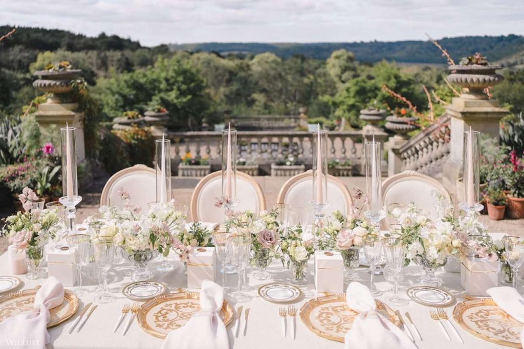 In questa foto un tavolo per matrimonio allestito sulla terrazza di un palazzo nobiliare. Sul tavolo sono presenti piatti di colore oro e bianco su cui sono poggiati tovaglioli di colore bianco. Al centro sono presenti composizioni di rose bianche e rose cappuccino alternate a portacandele di cristallo. Sullo sfondo si intravedono sedie classiche colore bianco e avorio e un giardino nobiliare