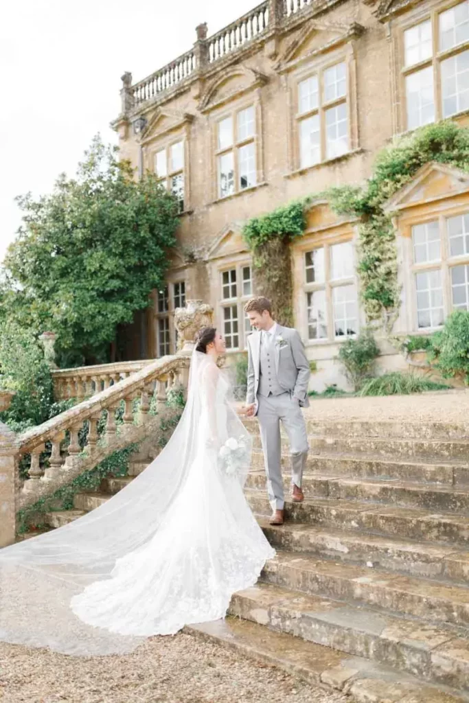 In questa foto due sposi si guardano sulle scale di una dimora storica per matrimonio in stile Bridgerton. La sposa indossa un abito semi sirena abbinato ad un velo lungo di tulle taglio laser