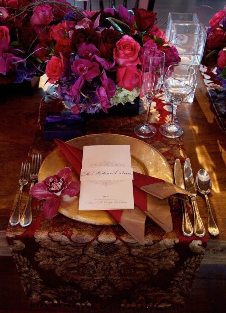 In questa foto una mise en place di matrimonio in stile barocco nei toni dell'oro, del viola e del fucsia. In primo piano è mostrato il piatto di colore oro con menù, tovagliolo abbinato e un orchidea come segnaposto. Al centro del tavolo è presente una composizione di rose e ciclamini