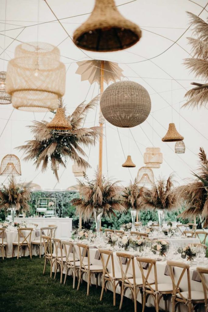 In questa foto un allestimento di matrimonio in stile Boho Chic realizzato su un prato. Sono presenti tavoli imperiali con sedie campagnole decorati con tovaglie di colore bianco, vasi di vetro trasparente con pampas e piccole composizioni di fior di colore rosa. Sui tavoli sono sospesi lampadari in paglia