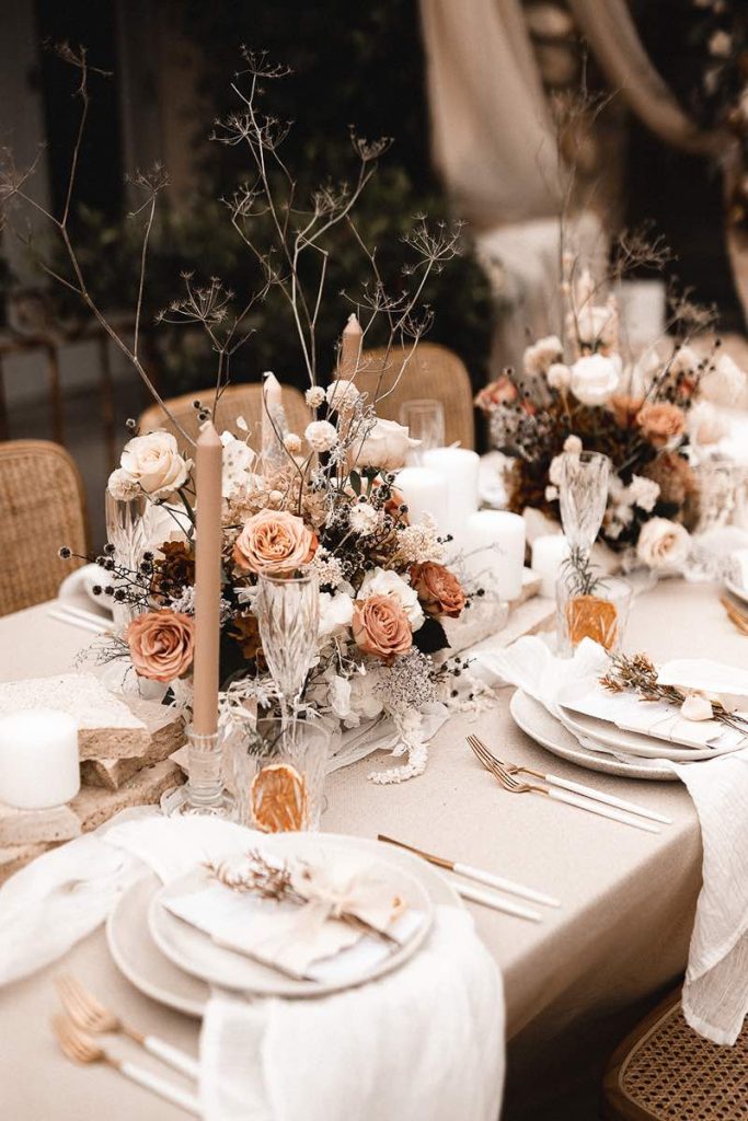 In questa foto un tavolo imperiale allestito per un matrimonio in stile boho chic con composizione di rose cappuccino e di colore bianco, candele beige in portacandele di vetro, tovagliato beige, posate dorate e di colore bianco