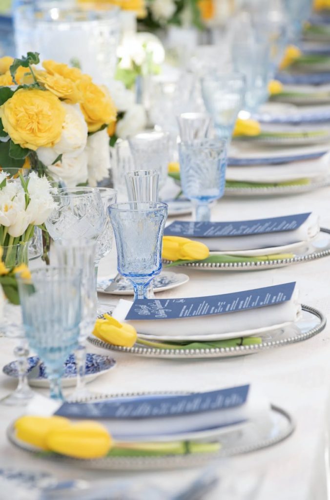 In questa foto un tavolo imperiale per matrimonio decorato in stile Capri con piatto da portata di vetro trasparente, menù di colore blu, tulipani di colore giallo, bicchieri di colore blu, piattino del pane in ceramica decorata con motivo maiolicato di colore blu