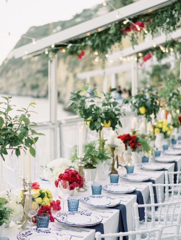 In questa foto un tavolo imperiale per matrimonio allestito in stile Capri con centrotavola di limoni, rose di colore giallo e rosso, bicchieri di colore blu, piatti di colore bianco e blu, tovaglia bianca e tovaglioli di colore blu
