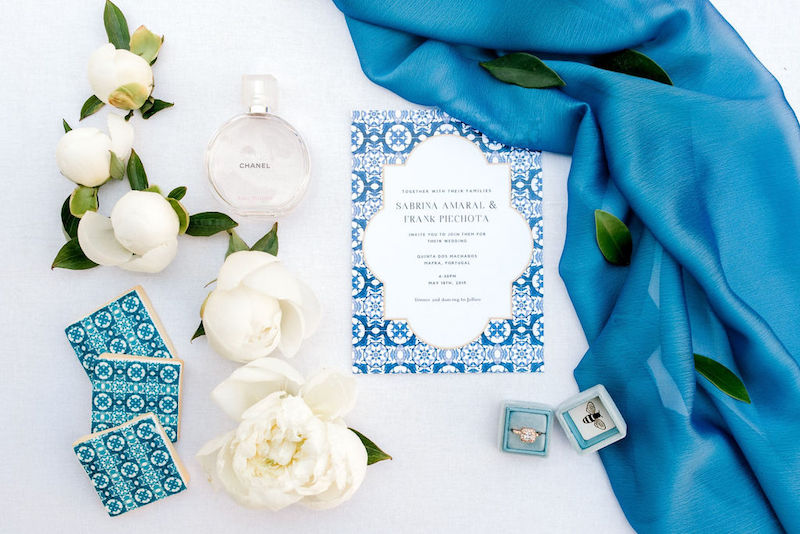 In questa foto un invito per matrimonio in stile Capri decorato con maioliche di colore blu. Attorno sono presenti maioliche, fiori di colore bianco, un anello con diamente e un drappo di seta di colore blu