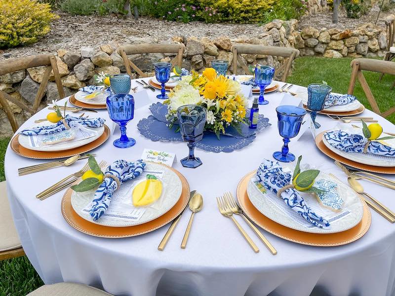 In questa foto una mise en place di matrimonio in stile Capri con tovaglia bianca, tovagliolo con motivo maiolicato blu e bianc, limoni e fiori di colore bianco e giallo, bicchieri di colore blu