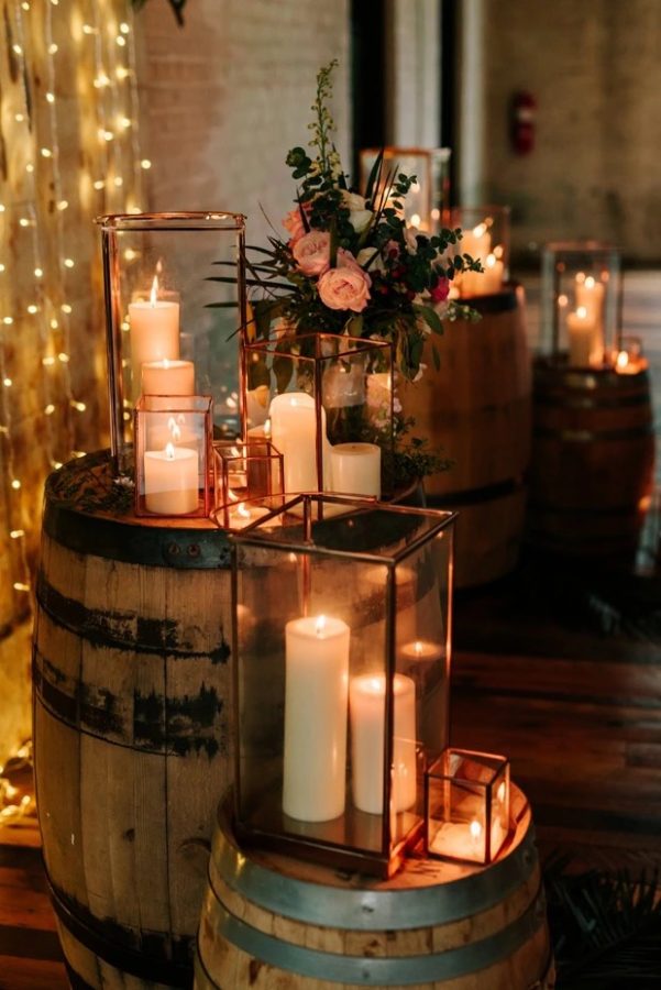 In questa foto addobbi di nozze in stile Country Chic composti da barili di legno, candele bianche, copricandele di vetro trasparente di forma rettangolare e fiori. Dietro si intravede anche un muro di piccole luci