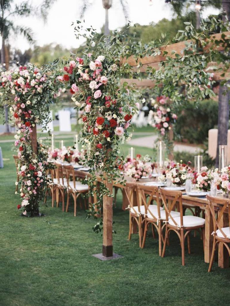 In questa foto un allestimento per matrimonio in stile coutry chic. Su una struttura in legno sono intrecciate rose di colore rosa e rosso. Le stesse composizioni sono presenti su un tavolo imperiale. Attorno al tavolo sono posizionate sedie campagnole