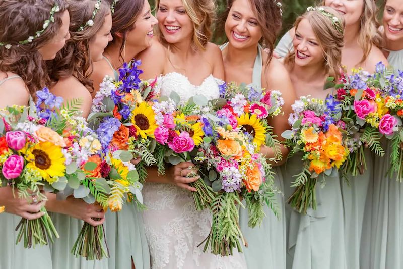 In questa foto una sposa con le sue damigelle che indossano abiti di colore verde salvia. Tutte tengono tra le mani bouquet in stile country chic composti da rose, tulipani, girasoli e lilium
