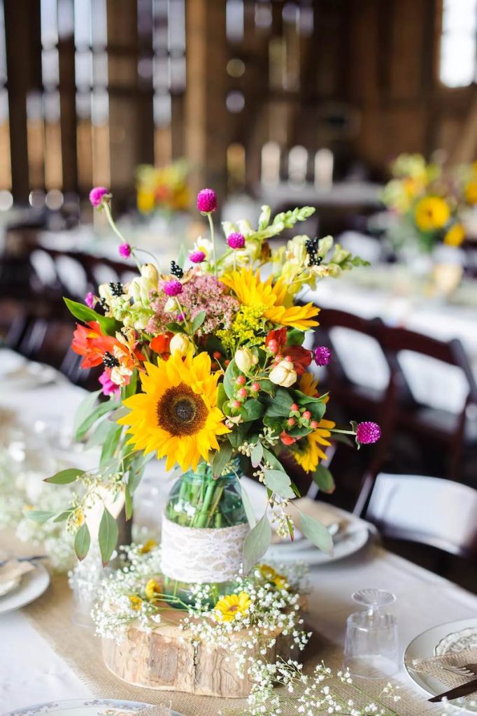 In questa foto un centrotavola per matrimonio in stile country chic con girasoli e fiori di campo di colore rosso e fucsia