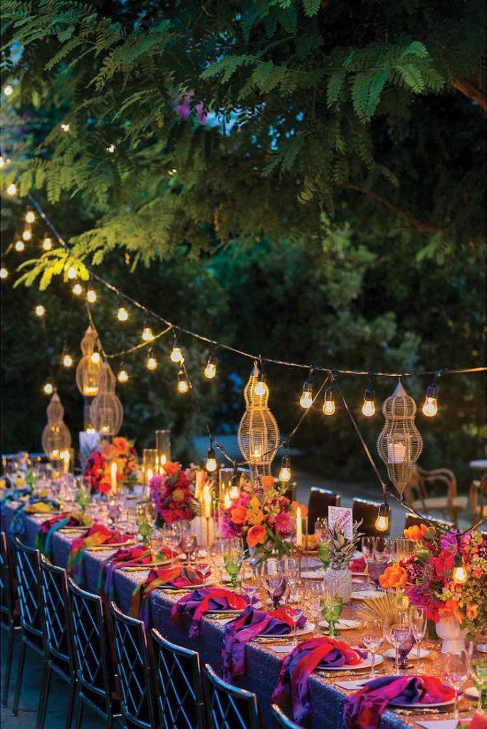 In questa foto un allestimento di matrimonio in stile etnico con tovaglie di colore rosa, tovaglioli di colore fucsia, fiori di colore arancione e fucsia, bicchieri di colore verde e viola, lampadari in paglia