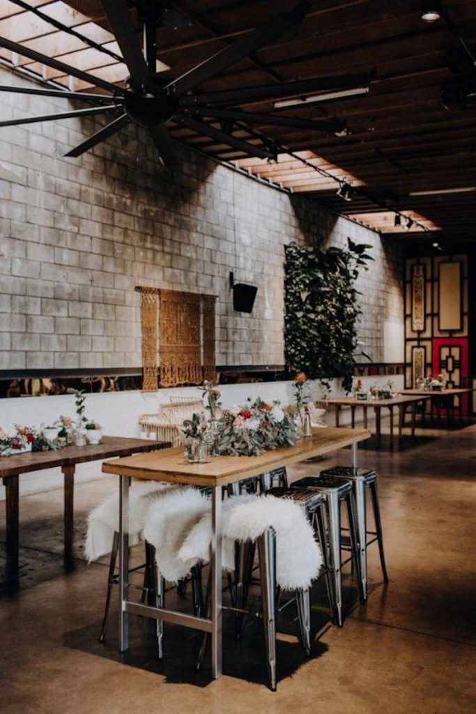 In questa foto un allestimento per matrimonio in stile industriale con tavoli in legno e acciaio alti abbinati a sgabelli in acciaio. I tavoli sono decorati con centrotavola con fiori di colore rosso, bianco e rosa e foglie di eucalipto