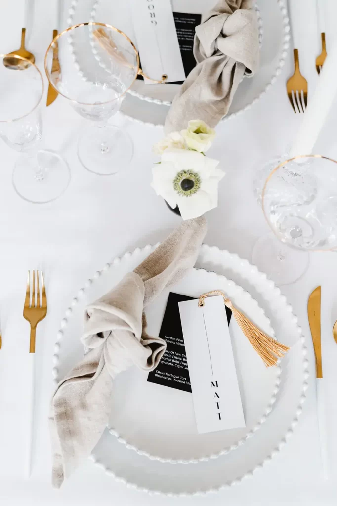 In questa foto una mise en place in. stile minimal con piatti bianchi, tovagliolo di colore beige, posate. di colore bianco e oro, fiori bianchi
