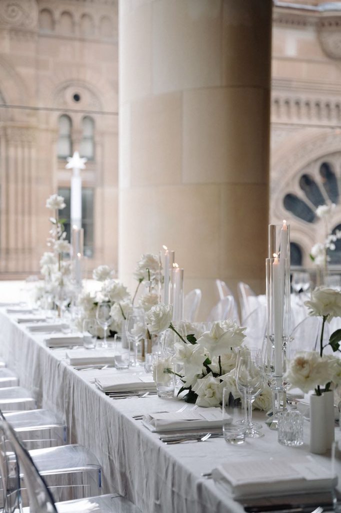 In questa foto un tavolo per matrimonio allestito in stile minimal con rose di colore bianco, vasetti di vetro trasparente e tovaglioli abbinati