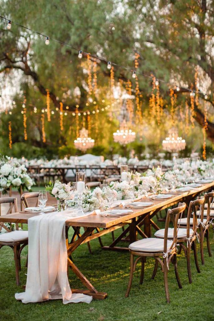 In questa foto un allestimento di matrimonio natural chic su un prato con luci pendenti dagli alberi, tavoli imperiali, fiori di colore rosa e bianco e runner di garza di colore beige