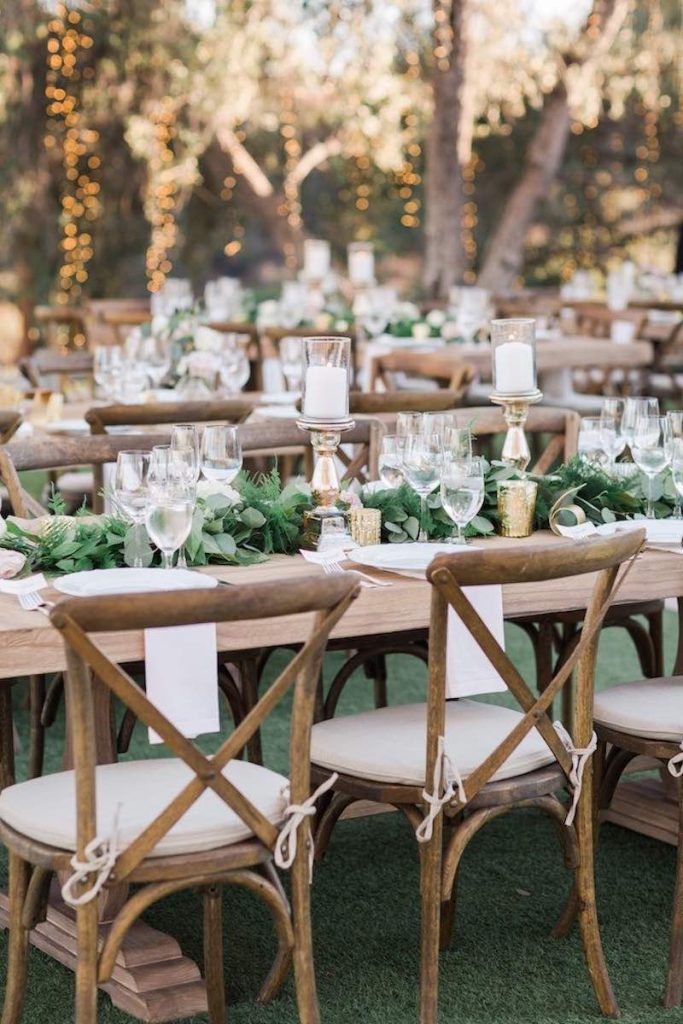 In questa foto tavoli per matrimonio natural chic in legno nudo decorati con runner di foglie verdi e portacandele di colore oro. Sono abbinate sedie campagnole con cuscini di colore beige