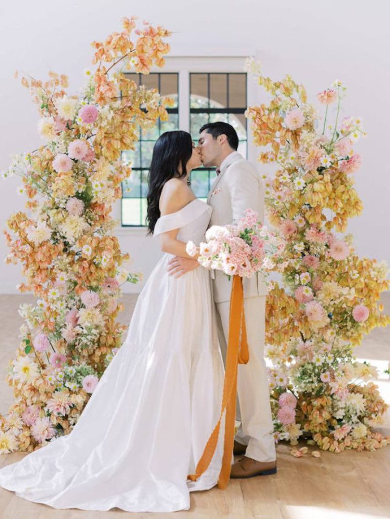 In questa foto due sposi si baciano davanti ad un arco di fiori per matrimonio romantico con rose di colore di rosa, fiori arancioni e bianchi e dalie