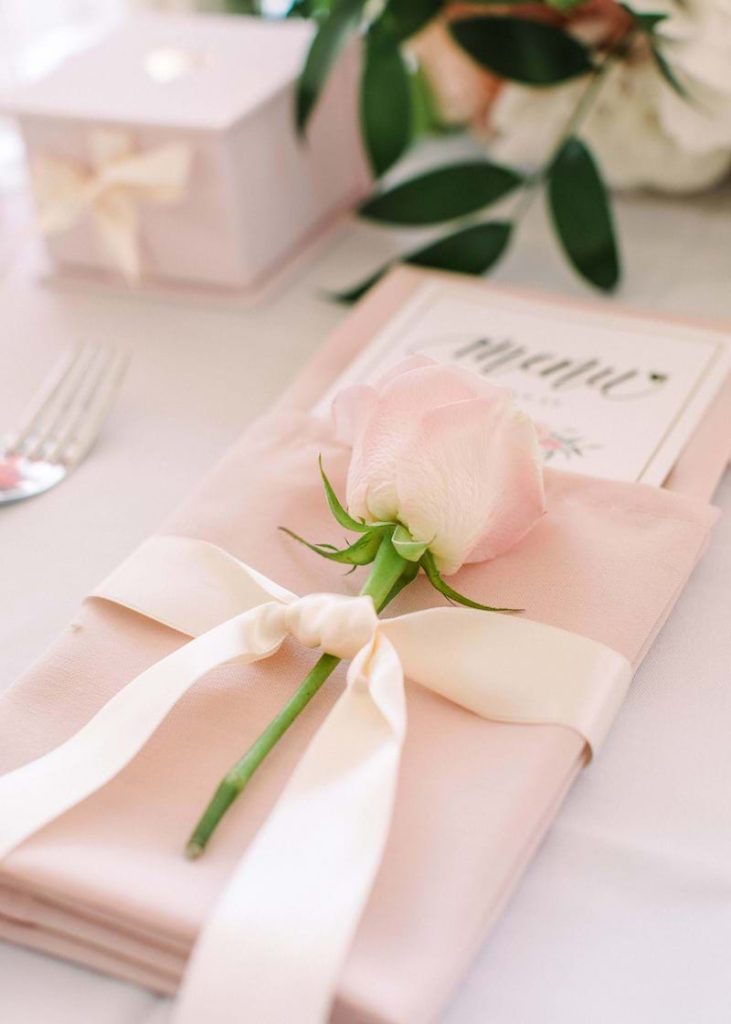In questa un segnaposto per matrimonio romantico realizzato con una rosa di colore legata ad un tovagliolo di stoffa di colore rosa con un nastro di stoffa di colore bianco. Dentro al tovagliolo è posizionato il menù
