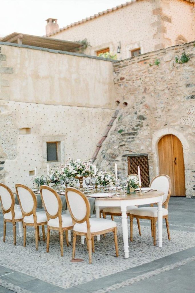 In questa foto un allestimento di un matrimonio in stile shabby chic in un antico casale con un tavolo ovale e sedie classiche, decorato con coppe con fiori di colore bianco e foglie e candele di colore bianco