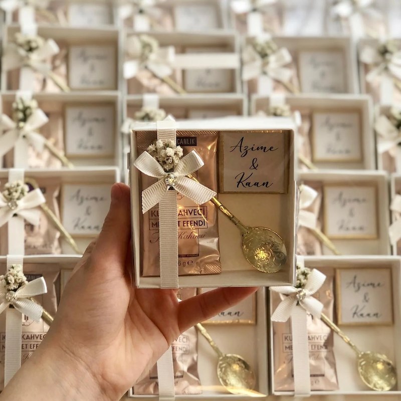 In questa foto bomboniere per matrimonio vintage composte da una scatoline di colore bianco, un pacchetto di confetti in confezione dorata, un cioccolatino con i nomi degli sposi e un cucchiaino dorato in stile retrò, Tutto è fermato da un nastro bianco con fiori secchi abbinati