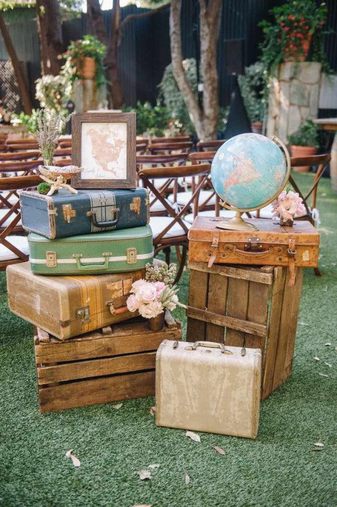 In questa foto decorazioni per matrimonio in stile vintage realizzati con valigie antiche, un mappamondo e una cartina posizionate su un prato dietro le sedie in legno per la cerimonia