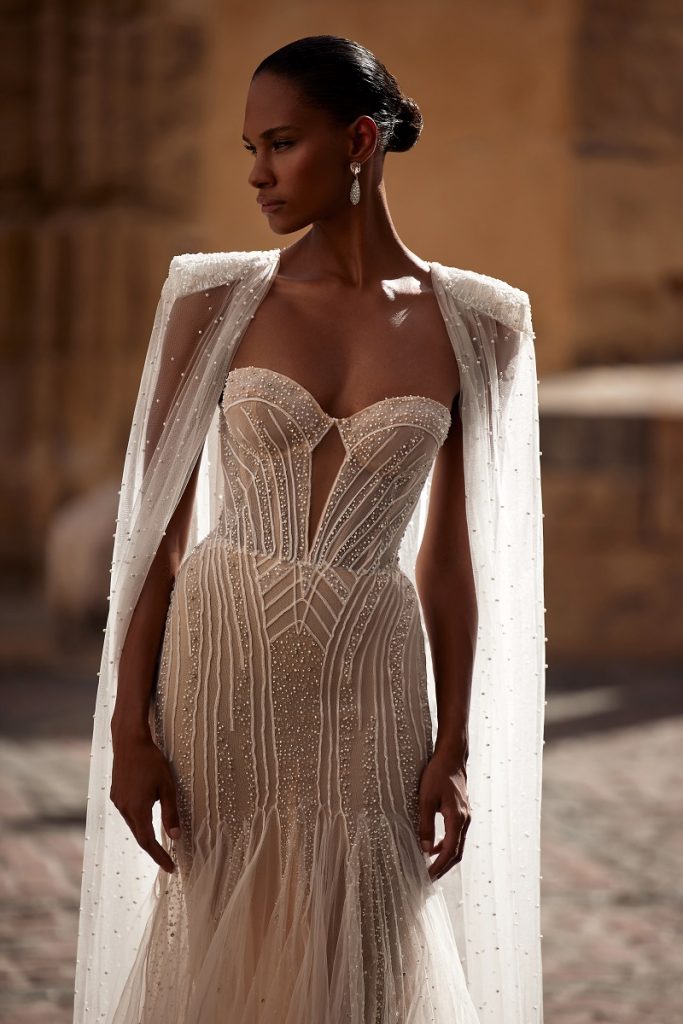 In questa foto una modella indossa un abito da sposa trasformabile 2023: la mantella poggiata sulle spalle è tempestata di micro perle. Fa parte della collezione Julie Vino 2023