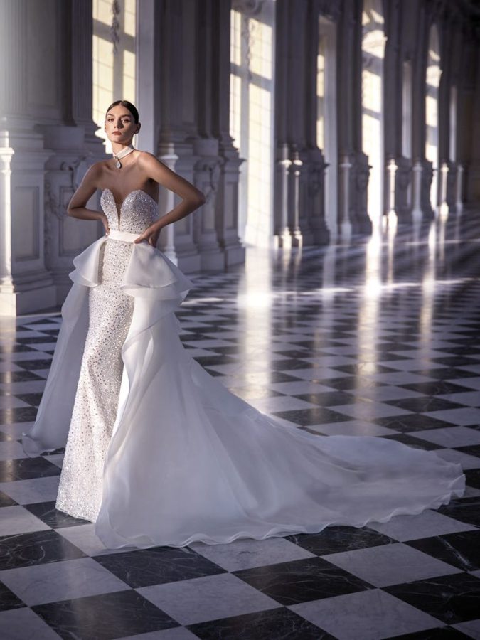 In questa foto una modella posa indossando un abito da sposa Pronovias 2023 con sopragonna removibile